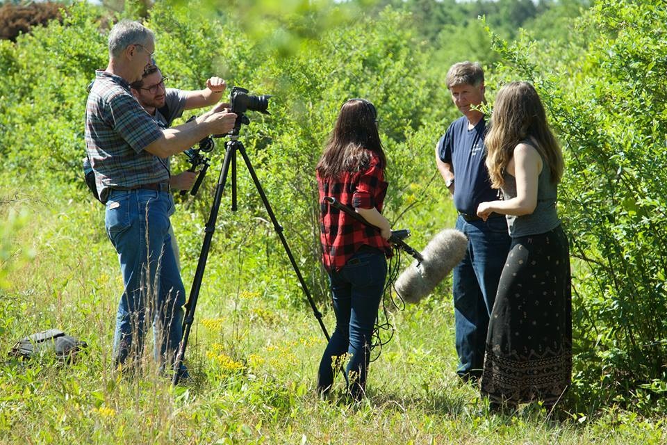 Filmmaking team capturing footage in a lush green setting, featuring a camera operator adjusting equipment, sound technician holding a microphone, and two individuals engaging in conversation, surrounded by vibrant foliage and wildflowers.