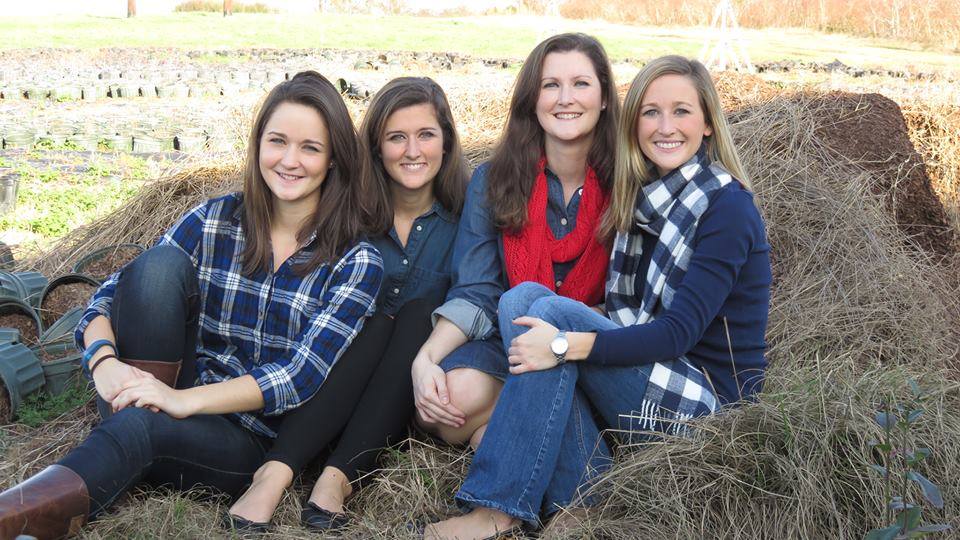 Four young women sitting together on a grassy area at a farm, dressed in casual outfits featuring plaid and denim. The background includes agricultural elements, such as planting containers and a field, capturing a cheerful and friendly atmosphere.